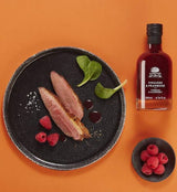 A L'Olivier Essig mit Himbeere neben rosa gebratener Entenbrust und frischen Himbeeren auf orangefarbenem Hintergrund – fruchtig-aromatische Essigspezialität, ideal für Wild- und Geflügelgerichte, Salate sowie süß-herzhafte Gourmet-Kreationen