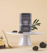 Schwarzes Trüffel-Olivenöl von A L'Olivier in eleganter Metalldose auf einem Podest präsentiert. Umgeben von schwarzen Trüffeln, Trüffelscheiben und Olivenzweigen zeigt die Szene die hochwertige Gourmetqualität des aromatisierten Olivenöls.