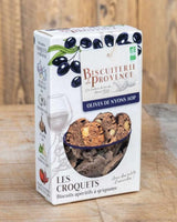 Verpackung von Biscuiterie de Provence „Les Croquets – Olives de Nyons AOP“: eine Schachtel mit Sichtfenster, gefüllt mit knusprigen, braunen Brotscheiben mit Mandeln und schwarzen Olivenstücken. Auf dem Design sind schwarze Oliven illustriert, dazu der Markenanhänger und Hinweise auf Bio-Qualität. Die Packung steht auf einem rustikalen Holztisch und wirkt wie ein hochwertiger Aperitif-Snack aus der Provence.