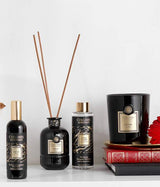 Collines de Provence Duftkollektion mit Raumspray Ebenholz, Duftdiffuser Ebenholz, Duftdiffuser-Nachfüllung Schwarze Rose und Duftkerze Zeder auf Regal mit Büchern und roter Blüte vor hellem Hintergrund.