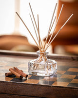 Durance Duftdiffuser Golden Teak Glasflakon mit Holzstäbchen auf Holztisch neben Zimtstangen warmes Wohnambiente