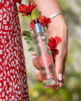 Durance Körperspray Pretty Poppy 100ml in transparenter Flasche wird in der Hand gehalten, umgeben von roten Blumen im Freien
