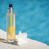 Durance Körperspray Sensual Monoi in transparenter Flasche am Pool mit Blüte in sommerlicher Szene
