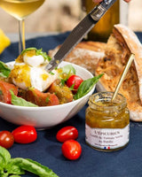 Serviervorschlag mit L'Épicurien Confit aus grünen Tomaten mit Basilikum zu Burrata, Tomaten, Basilikum und Brot, daneben ein geöffnetes Glas mit Löffel.