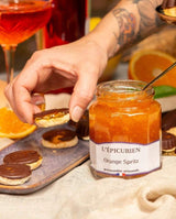 Serviervorschlag mit L'Épicurien Orange Spritz als Fruchtaufstrich auf Gebäck, daneben ein geöffnetes Glas mit Löffel sowie Orangenscheiben und ein Cocktail.