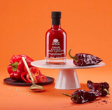 A L'Olivier Essig mit Paprika und Piment d'Espelette auf orangefarbenem Hintergrund mit frischer roter Paprika und getrockneten Espelette-Chilis – aromatisch-würzige Essigspezialität mit fruchtiger Paprikanote und feiner Schärfe für Grillgerichte, Salate, Gemüse und mediterrane Küche