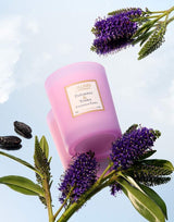Collines de Provence Duftkerze Patchouli & Tonkabohne in rosa Glas, dekorativ arrangiert mit violetten Blüten und Tonkabohnen