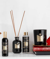Collines de Provence Duftkollektion mit Raumspray Ebenholz, Duftdiffuser Ebenholz, Duftdiffuser-Nachfüllung Schwarze Rose und Duftkerze Zeder auf Regal mit Büchern und roter Blüte vor hellem Hintergrund.