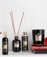 Collines de Provence Duftkollektion mit Raumspray Ebenholz, Duftdiffuser Ebenholz, Duftdiffuser-Nachfüllung Schwarze Rose und Duftkerze Zeder auf Regal mit Büchern und roter Blüte vor hellem Hintergrund.