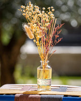 Durance Floral Reed Diffuser Fleur de Monoï Glasflakon mit Duftstäbchen und Trockenblumen auf Tisch im Garten