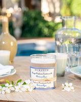 Durance Duftkerze Riviera Azur in dekorativem Glas auf einem Sommertisch im Freien, umgeben von weißen Blüten