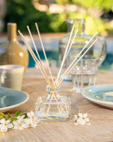 Durance Raumduft Diffuser Riviera Azur mit dekorativem Glasflakon und Duftstäbchen auf stilvoll gedecktem Tisch mit weißen Blüten, Geschirr und sommerlicher Outdoor-Dekoration