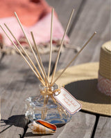 Durance Raumduft Diffuser Sonnenbad 100ml mit Duftstäbchen und dekorativem Glasflakon in sommerlichem Ambiente auf Holztisch mit Muschel und Sonnenhut