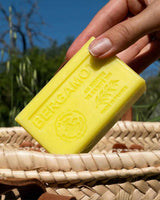 La Maison du Savon de Marseille Seife Bergamotte mit Bio-Sheabutter in einer Hand über einem Flechtkorb im natürlichen Außenbereich.