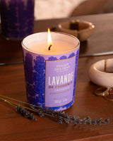 Panier des Sens Duftkerze Lavendel aus der Provence 180g im dekorativen Kerzenglas mit brennender Flamme auf Holztisch und Lavendelzweigen in stimmungsvollem Wohnambiente