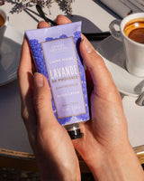 Panier des Sens Handcreme Lavendel 75ml in den Händen einer Person auf einem sonnigen Tisch mit Kaffeetassen. 